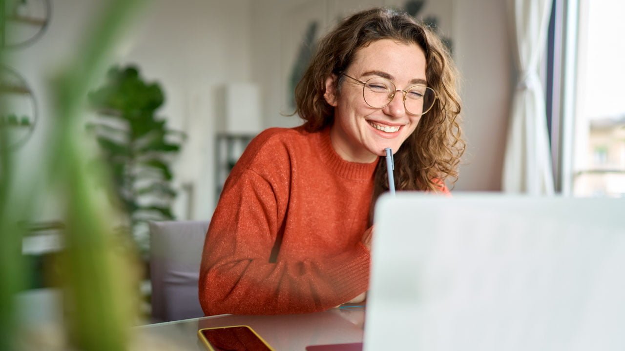 Young happy woman student using laptop watching webinar writing at home.